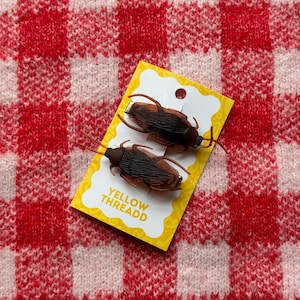 Peut inclure: Deux jouets en plastique en forme de cafards bruns et noirs sur une étiquette jaune avec le texte "YELLOW THREADD". L'étiquette est sur un fond de tissu à carreaux rouges et blancs.