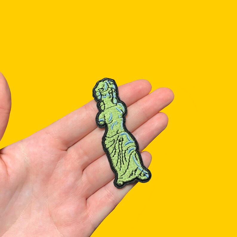 Puede incluir: Un parche bordado que representa una estatua verde de una mujer con un contorno azul.