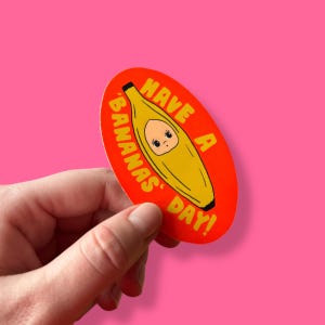 May include: Oval sticker with a yellow banana character inside a red peel, with the text "Have a Bananas Day!" in white. The sticker is held against a pink background.