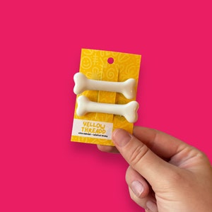 May include: Two white bone-shaped hair clips on a yellow card with the text "YELLOW THREADD". The card has a smiley face and doodle pattern. The background is a bright pink.