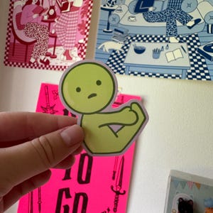 May include: A light green sticker featuring a cartoon character flexing its arm. The character has a round head with a simple face. The sticker is held in front of a pink poster with the words "TO GO" and illustrations of swords.