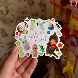 May include: A sticker featuring various cartoon characters, including a dog, Snoopy, a bunny, and a Monchhichi doll. The sticker has the text "ask me about my trinkets" and is decorated with hearts and stars.