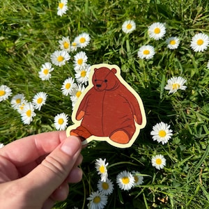 May include: A brown bear sticker with orange accents, held against a backdrop of green grass and white daisies. The sticker features a cartoon-style illustration of a seated bear with a friendly expression.