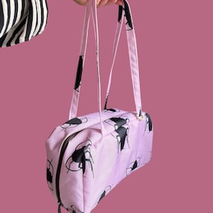 May include: A light pink rectangular bag featuring a repeating black graphic print of insects. The bag has two long shoulder straps, a dark brown zipper closure, and a small, round, bright yellow tag attached to the zipper pull.