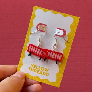 May include: Red barrier earrings with black supports, suspended from silver hooks. The earrings are presented on a yellow card with the text "YELLOW THREADD" against a red backdrop.