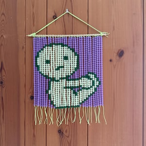 May include: A beaded wall hanging featuring a whimsical figure. The background is composed of small, purple beads, while the figure is created with white and green beads. The piece is suspended from a wooden dowel with a lime green cord and fringe.