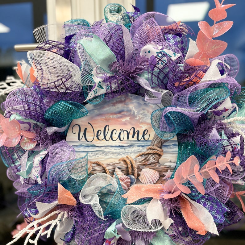 May include: A decorative wreath featuring a beach-themed design. The wreath incorporates shades of purple, teal, and white mesh, accented with coral-colored floral elements. A central wooden plaque displays the word "Welcome" with a beach scene.