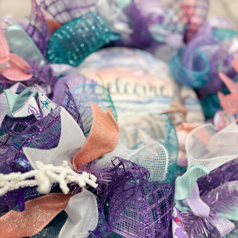 May include: A decorative wreath with a coastal theme. It features layers of purple, teal, and white mesh, accented with coral-colored ribbons and a central element with the word "welcome" over a beach scene.