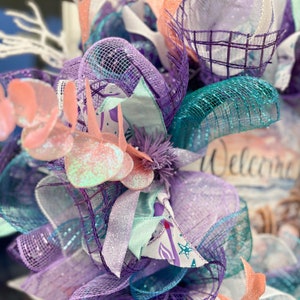 May include: A decorative wreath with a coastal theme, featuring layers of purple, teal, and coral mesh ribbons. Accented with glittery embellishments and the word "Welcome" in script.