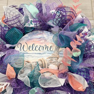 May include: A decorative wreath with a coastal theme. The wreath features a central sign with the word "Welcome" and a beach scene. It is adorned with teal, purple, and white mesh, along with seashell accents and coral-colored embellishments. The wreath is designed to be hung on a door or wall.