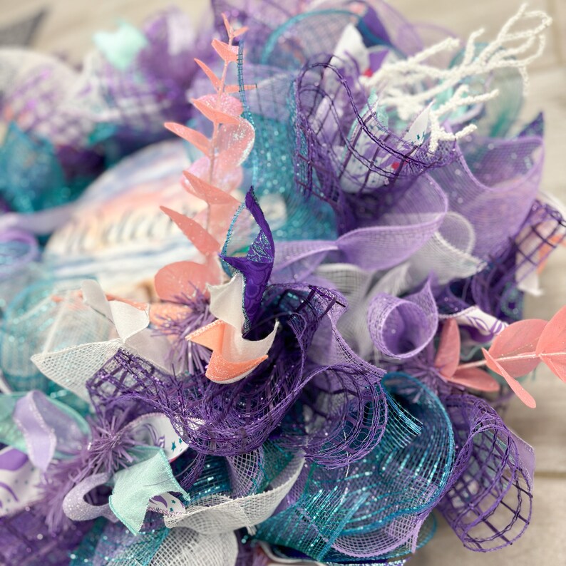 May include: A close-up of a decorative wreath featuring layers of purple, teal, and white mesh. The wreath is adorned with coral-colored and white decorative elements, creating a textured and colorful design. The wreath is suitable for home decor.