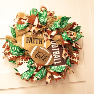 Op de afbeelding: Een groene, bruine en gouden voetbal-krans met jute, linten en houten voetballen met de woorden "Faith", "Fall" en "Football".