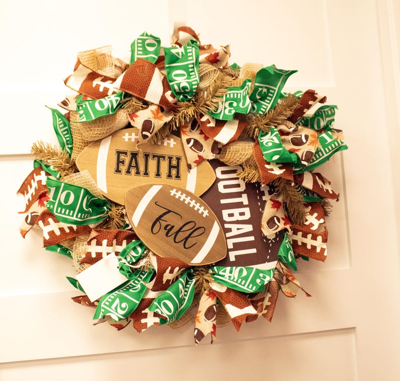 Op de afbeelding: Een groene, bruine en gouden voetbal-krans met jute, linten en houten voetballen met de woorden "Faith", "Fall" en "Football".