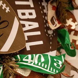 Op de afbeelding: Een bruine en witte voetbalvormige krans met het woord "Football" erop gedrukt. De krans is versierd met bruine, witte en groene stofresten, waaronder een groene strook stof met de nummers "40 3" erop gedrukt.