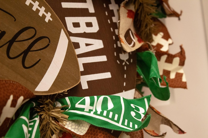 Op de afbeelding: Een bruine en witte voetbalvormige krans met het woord "Football" erop gedrukt. De krans is versierd met bruine, witte en groene stofresten, waaronder een groene strook stof met de nummers "40 3" erop gedrukt.