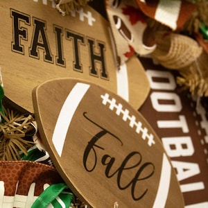 Op de afbeelding: Houten voetbalschilderingen met de woorden "Faith" en "Fall" in zwarte schrijfletters. De schilderingen zijn omgeven door bruin en groen stof met voetbalpatronen.