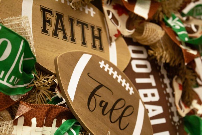 Op de afbeelding: Houten voetbalschilderingen met de woorden "Faith" en "Fall" in zwarte schrijfletters. De schilderingen zijn omgeven door bruin en groen stof met voetbalpatronen.