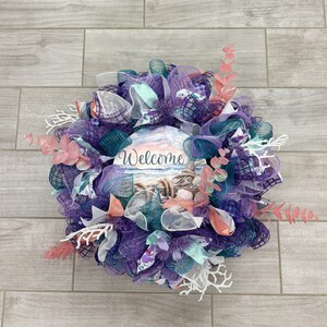 May include: A decorative wreath with a beach theme. The wreath features a central sign with the word "Welcome" and a beach scene. It is made of purple, teal, and white mesh, with coral and seashell accents. The wreath is approximately 18 inches in diameter.