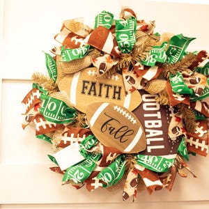 Op de afbeelding: Een bruine en groene voetbalthema krans met jute, linten en houten voetballen met de woorden "Faith" en "Fall" erop.