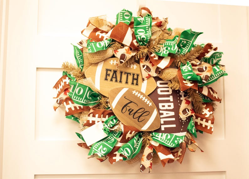Op de afbeelding: Een bruine en groene voetbalthema krans met jute, linten en houten voetballen met de woorden "Faith" en "Fall" erop.