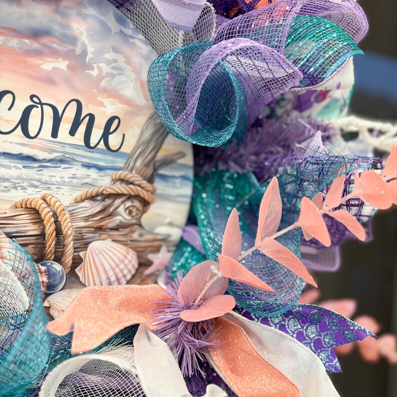 May include: A coastal-themed wreath with the word "Welcome" visible. The wreath features a watercolor image of a beach scene, seashells, and decorative mesh in shades of teal, purple, and coral. Rope and faux foliage add texture to the design.