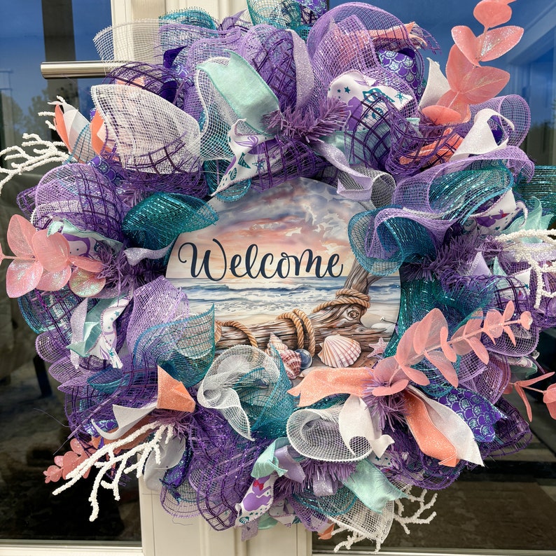 May include: A decorative wreath with a coastal theme. It features a central sign with the word "Welcome" and a beach scene. The wreath is adorned with purple, teal, and white mesh, ribbons, and faux coral and foliage. Shells and rope add to the nautical design.