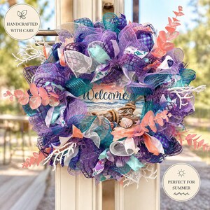 May include: A handcrafted, colorful wreath with a coastal theme. It features shades of purple, teal, and white mesh, accented with faux coral, seashells, and a wooden sign that reads "Welcome". Perfect for summer.