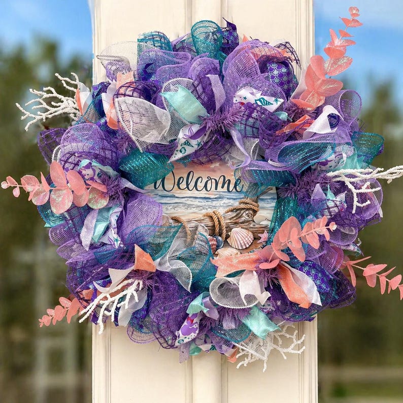 May include: A decorative wreath with a coastal theme. It features a mix of purple, teal, and white mesh, accented with coral-colored leaves and white branches. A central sign reads "Welcome" above a beach scene with seashells.