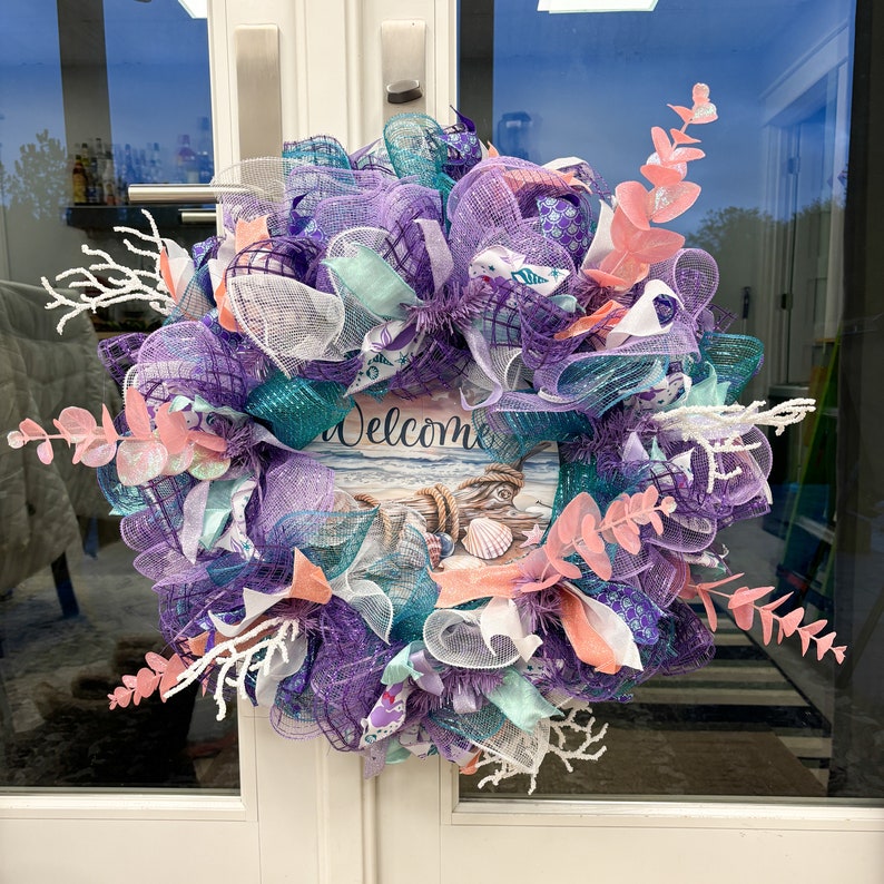 May include: A decorative wreath with a coastal theme. It features a base of purple, teal, and white mesh, accented with coral and white decorative elements. A sign in the center reads "Welcome" with a beach scene.