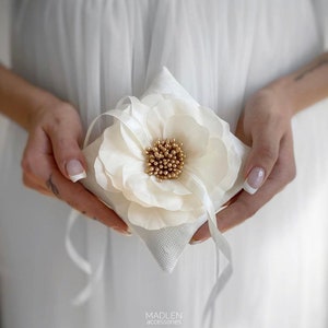 May include: White satin ring pillow with a large white flower and gold accents. The pillow has a ribbon tied around it.
