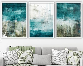 May include: Three framed abstract art prints with teal, white, and brown brushstrokes. The artwork is displayed above a gray sofa with decorative pillows and a green throw blanket. A white coffee table with a cup and saucer is in front of the sofa.