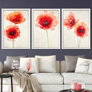 Puede incluir: Tres impresiones enmarcadas en acuarela de amapolas rojas. Cada impresión presenta una disposición diferente de las flores, con tallos verdes y un fondo crema. Los marcos son blancos y la obra de arte se exhibe sobre un sofá blanco.