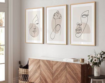 May include: Three minimalist line art prints with abstract faces in black on a beige background. The prints are framed in gold and hung on a white wall above a wooden cabinet.