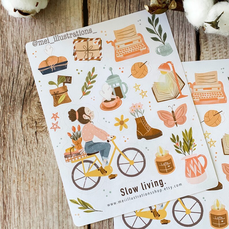 May include: Two sticker sheets with illustrations of a woman riding a yellow bicycle, a typewriter, coffee, books, and other cozy items. The text "Slow living" is at the bottom.
