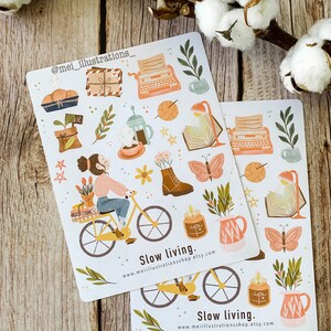 May include: Two sticker sheets with illustrations of a person riding a bicycle, a typewriter, books, plants, and other cozy items. The text "Slow living" and a website address are printed on the stickers. The stickers have a soft, pastel color palette.