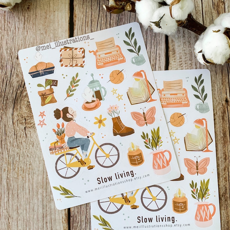 May include: Two sticker sheets with illustrations of a person riding a bicycle, a typewriter, books, plants, and other cozy items. The text "Slow living" and a website address are printed on the stickers. The stickers have a soft, pastel color palette.