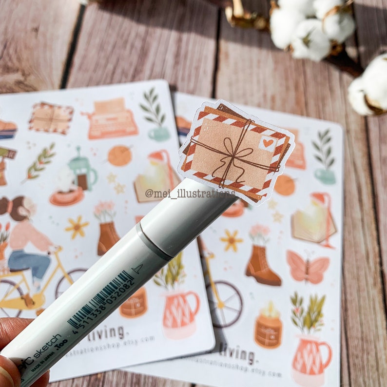 May include: A white Copic sketch marker rests on a sheet of stickers. The stickers feature illustrations of envelopes, flowers, and other stationery items in shades of brown, orange, and green. The sticker sheet has the text "living" and "mei_illustrations".