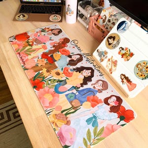 May include: A floral mousepad featuring illustrations of women among flowers, with the text "@mei_illustrations". A laptop, water bottle, and stickers are also on the wooden desk.