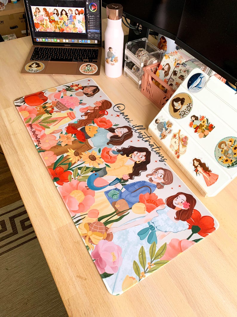 May include: A floral mousepad featuring illustrations of women among flowers, with the text "@mei_illustrations". A laptop, water bottle, and stickers are also on the wooden desk.