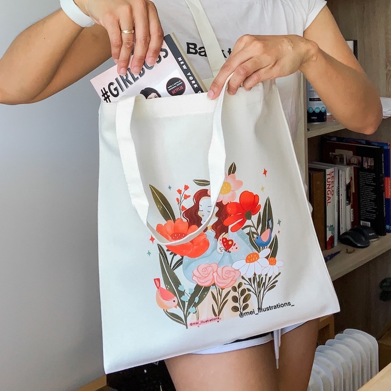 May include: A cream-coloured tote bag featuring a vibrant illustration of a woman amidst flowers and birds. The bag has long straps, and a book with "#GIRLBOSS" is visible inside. The artist's name, "@mei_illustrations_," is printed on the bag.