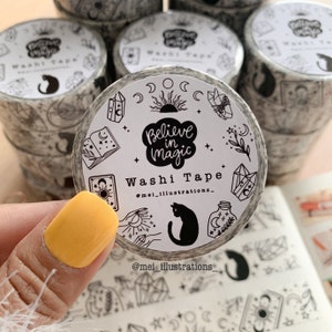 May include: A roll of washi tape with a black and white design featuring the text "Believe in Magic" and illustrations of crystals, moons, and a black cat. The tape is held by a hand with a yellow nail.
