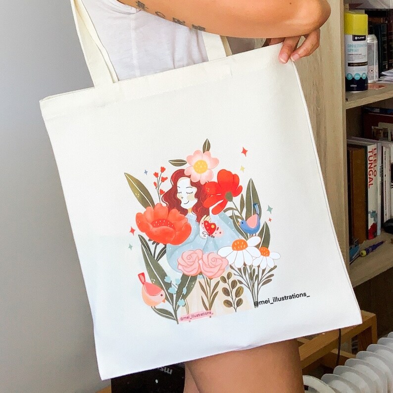 May include: Off-white tote bag with a vibrant illustration of a woman amidst flowers and birds. The design includes red poppies, pink roses, and daisies. The artist's signature, "mei_illustrations_," is printed at the bottom of the bag.