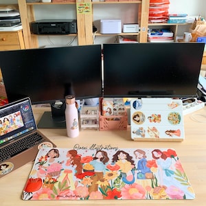 May include: A desk setup featuring two black computer monitors, a laptop, and a floral-themed mousepad. A pink water bottle and various desk organizers are also visible. The mousepad has a colorful illustration of women and flowers.