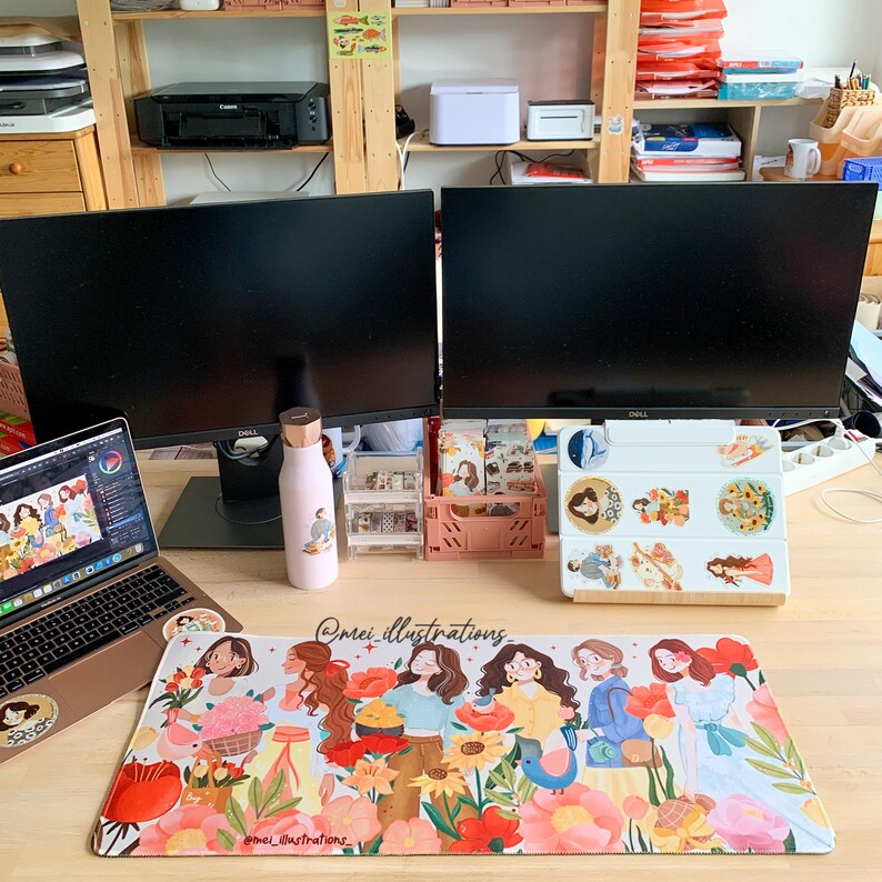 May include: A desk setup featuring two black computer monitors, a laptop, and a floral-themed mousepad. A pink water bottle and various desk organizers are also visible. The mousepad has a colorful illustration of women and flowers.