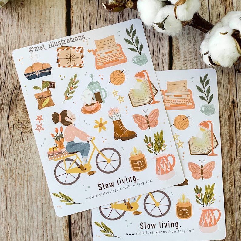 May include: Two sticker sheets with illustrations of a woman on a bicycle, a typewriter, coffee, books, and butterflies. The text "Slow living" and a website address are printed on the stickers. The stickers have a soft, pastel color palette.