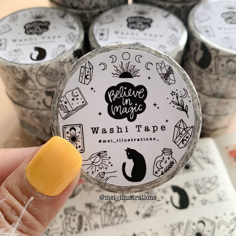 May include: A roll of washi tape with a black and white design. The tape features the text "Believe in Magic" and illustrations of crystals, moons, stars, and a black cat. The brand name "@mei_illustrations_" is also visible.