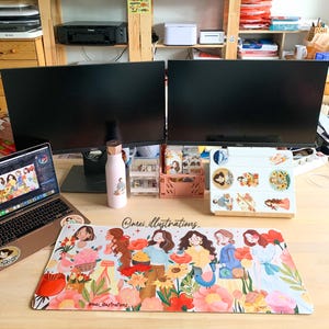 May include: A desk setup featuring a laptop, two large monitors, and a colorful mousepad. The mousepad displays an illustration of women with flowers. A pink water bottle and various desk organizers are also visible.