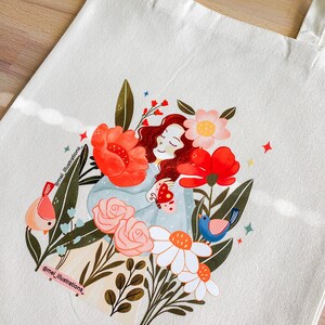 May include: Cream tote bag with a vibrant illustration of a person amidst flowers and birds. The design showcases red, pink, and white blooms, with a blue and pink bird on each side. The artist's name, @mei_illustrations_, is printed on the bag, adding a personal touch.