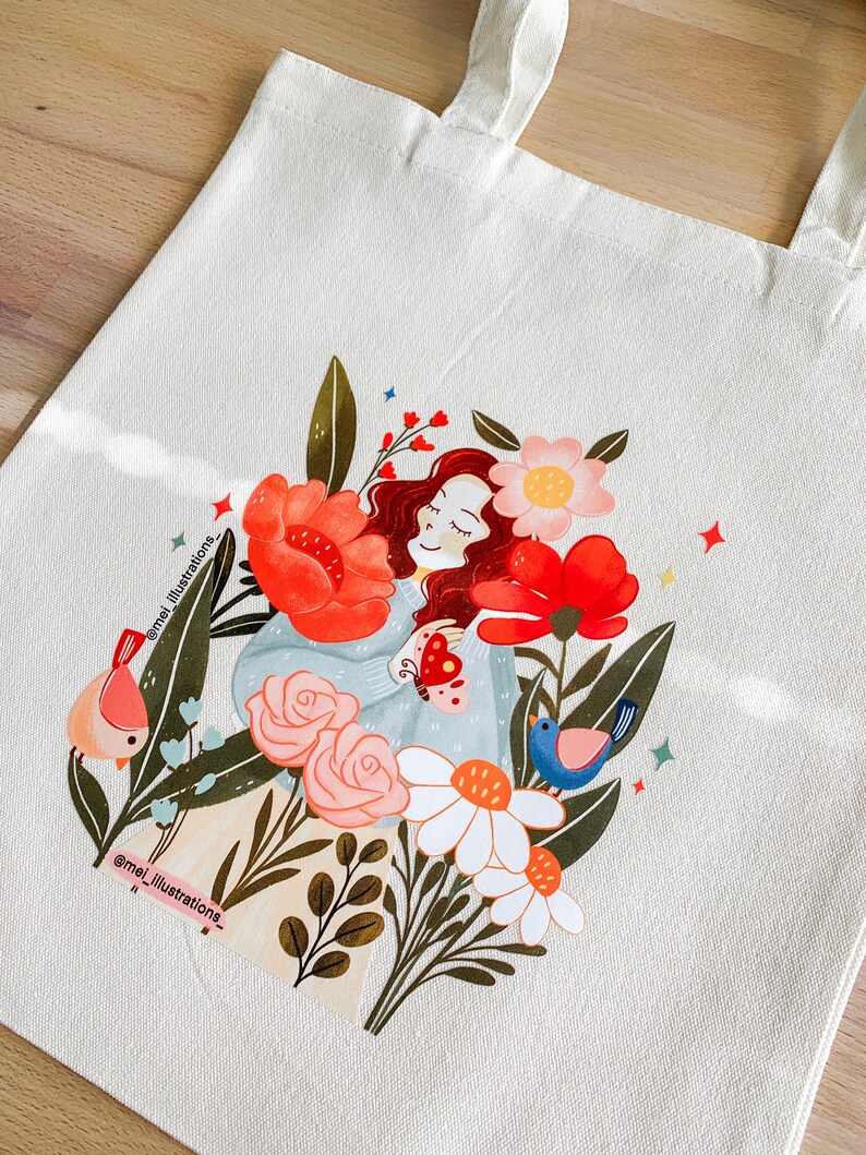 May include: Cream tote bag with a vibrant illustration of a person amidst flowers and birds. The design showcases red, pink, and white blooms, with a blue and pink bird on each side. The artist's name, @mei_illustrations_, is printed on the bag, adding a personal touch.