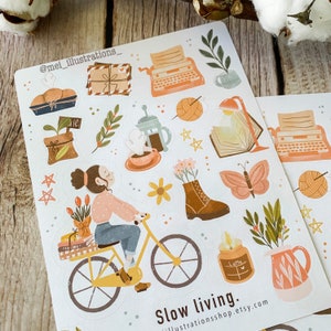 May include: A sheet of stickers with a "Slow living" theme. The stickers feature illustrations of a woman on a bicycle, a typewriter, coffee, books, flowers, and a butterfly. The color palette includes shades of orange, brown, and green.